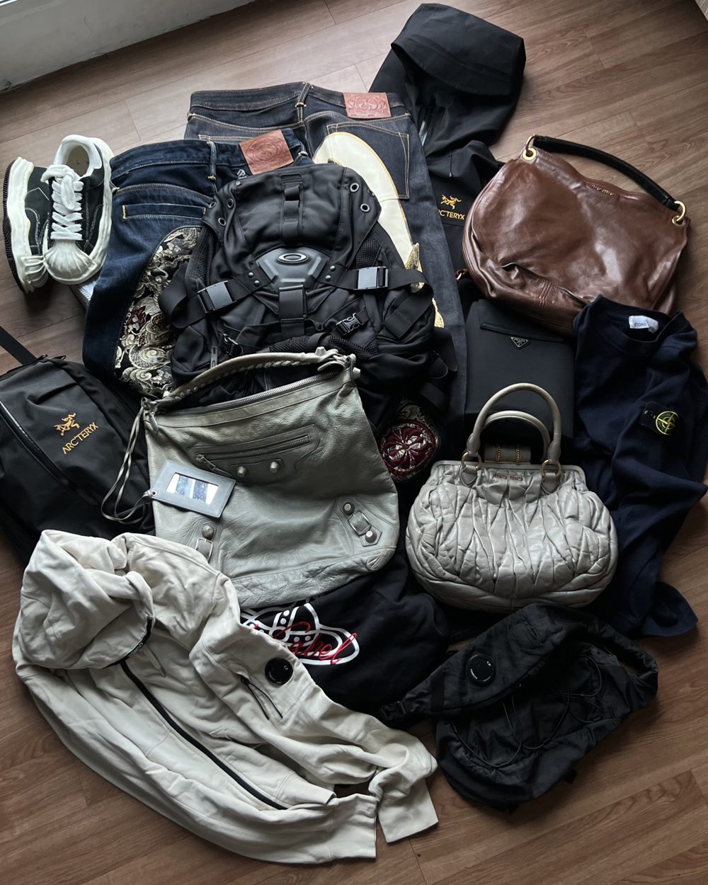 A sourcing haul laid out on a wooden floor: Balenciaga, Miu Miu and Prada bags, an Oakley backpack, Arc’teryx, Stone Island and C.P. Company pieces, denim and sneakers